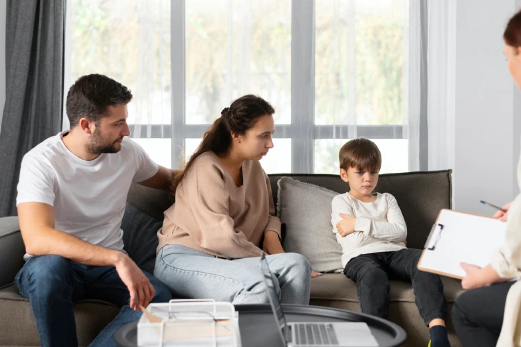 A family therapy session with a concerned couple and their upset child sitting on a sofa, facing a therapist holding a clipboard. Natural light filters through large windows.