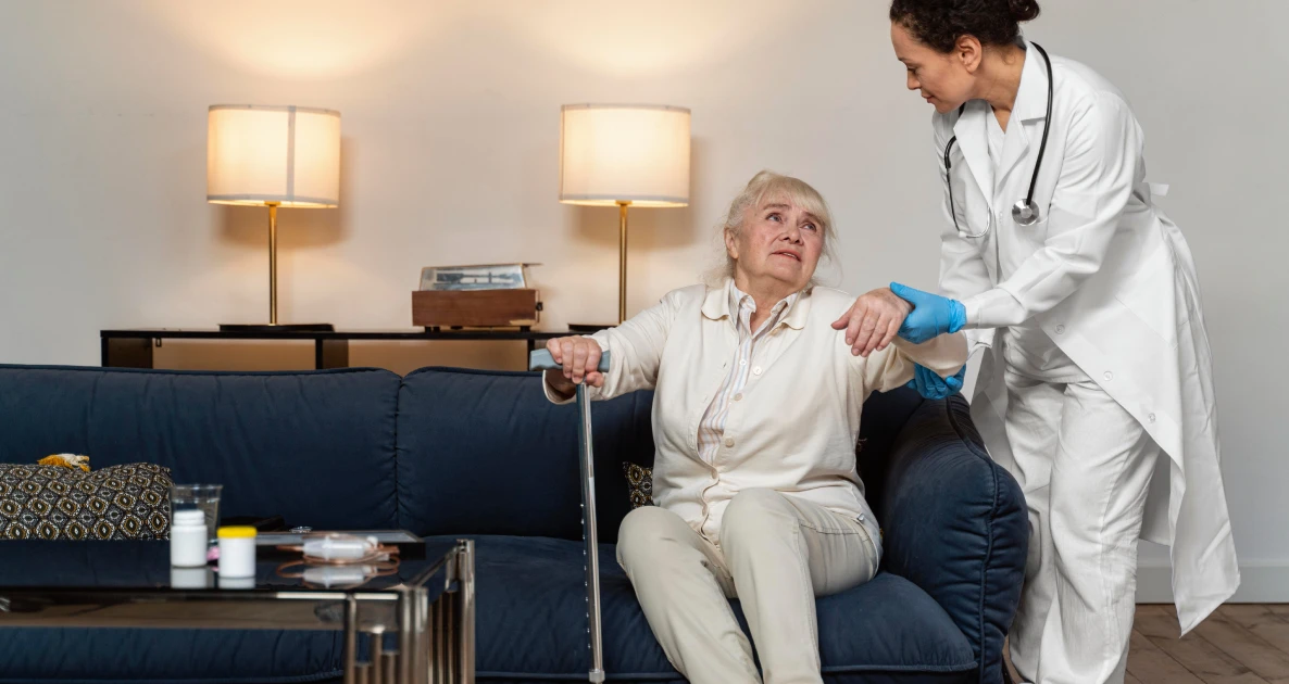 Caregiver helps an elderly woman with a cane in a warm living room.