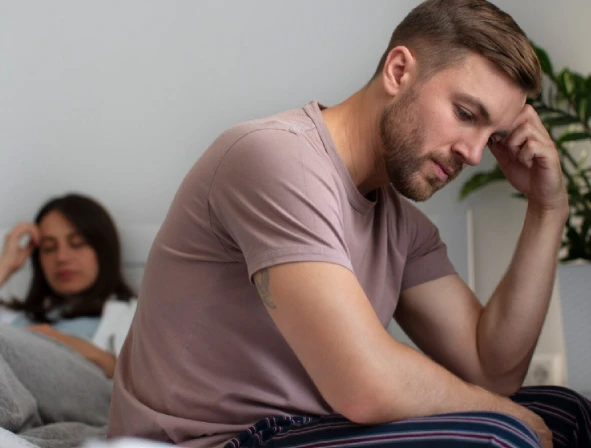 Man on bed looks concerned; woman sits blurred in background.