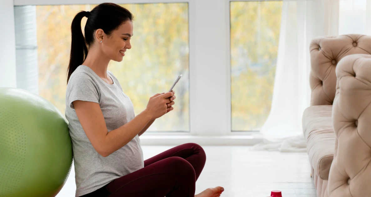 Pregnant woman smiles at phone beside yoga ball in bright room.