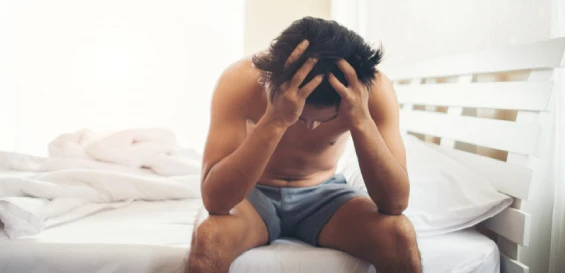 Shirtless man sits on bed, head in hands, distressed.