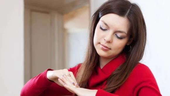 A woman in a red sweater applies lotion to her hands indoors. She appears focused and calm, with a neutral background enhancing the serene mood.