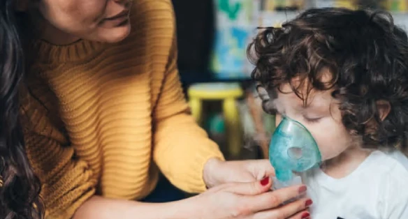 A woman in a yellow sweater assists a young child with curly hair using a nebulizer mask. The setting appears caring and attentive.