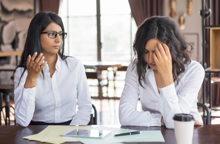 Two women in an office argue; one gestures, the other looks stressed.