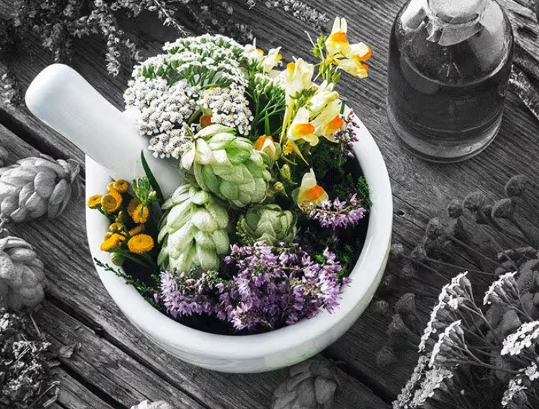 A white mortar filled with vibrant herbs and flowers, including hops, white blossoms, purple blooms, and yellow accents, rests on rustic wooden boards.