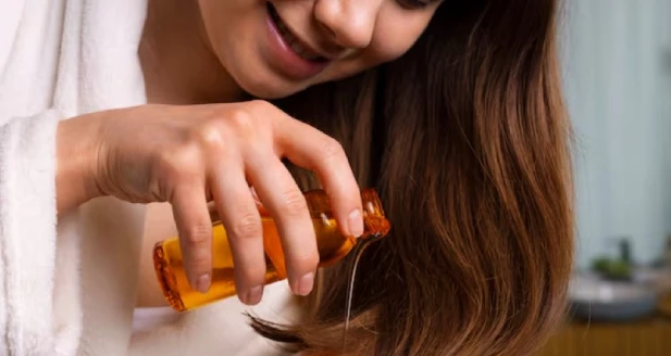 A young woman in a white robe smiles while pouring oil from a small amber bottle onto her long, brown hair, suggesting a self-care or hair treatment routine.