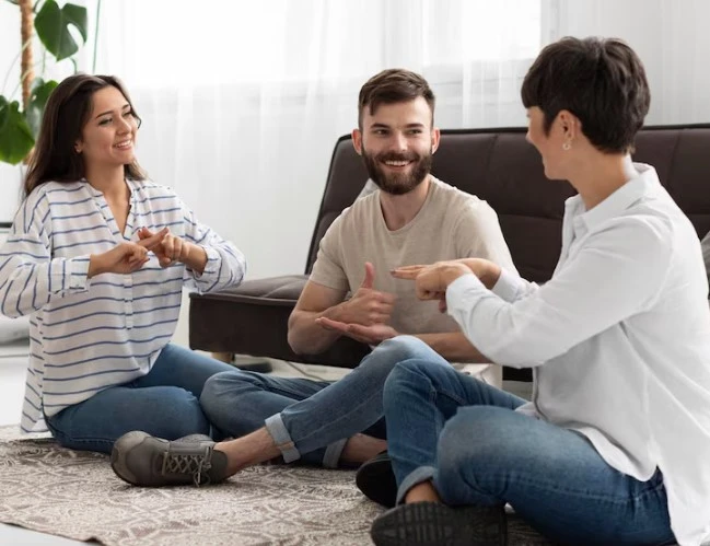 Three people sit smiling, using sign language in sunlight.