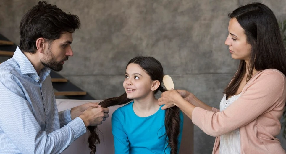 Man and woman braid a smiling girl’s hair on a sofa.