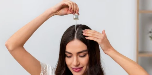 A woman with long brown hair is smiling as she applies hair serum to her scalp with a dropper. The background is soft and neutral, creating a calming tone.