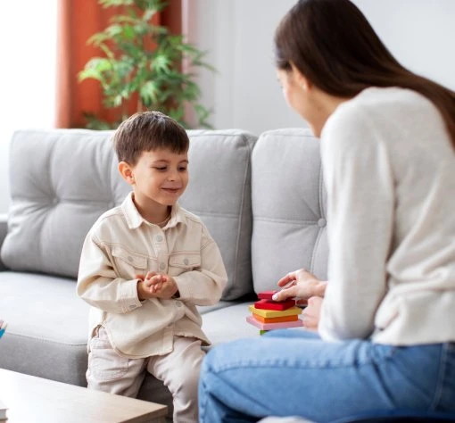 Boy smiles on sofa as woman stacks blocks in cozy, playful scene.