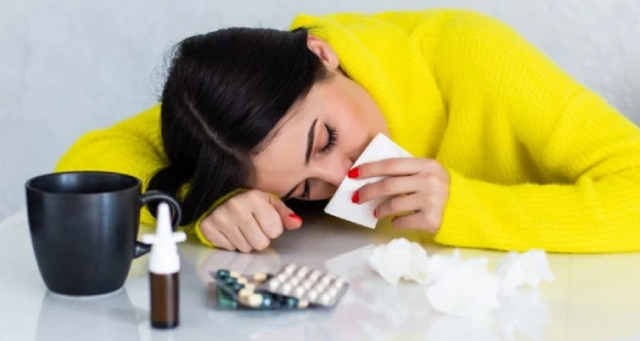 Woman in yellow sweater rests on table with tissue, surrounded by mug, meds, spray, and used tissues.