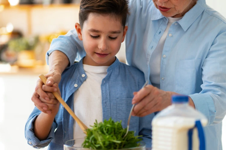 A young boy and an older adult in a kitchen, both wearing blue shirts, share a bonding moment while mixing a salad. The atmosphere is warm and nurturing.