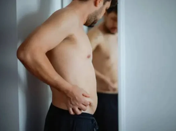 Shirtless man checks his waist in a mirror, looking thoughtful.