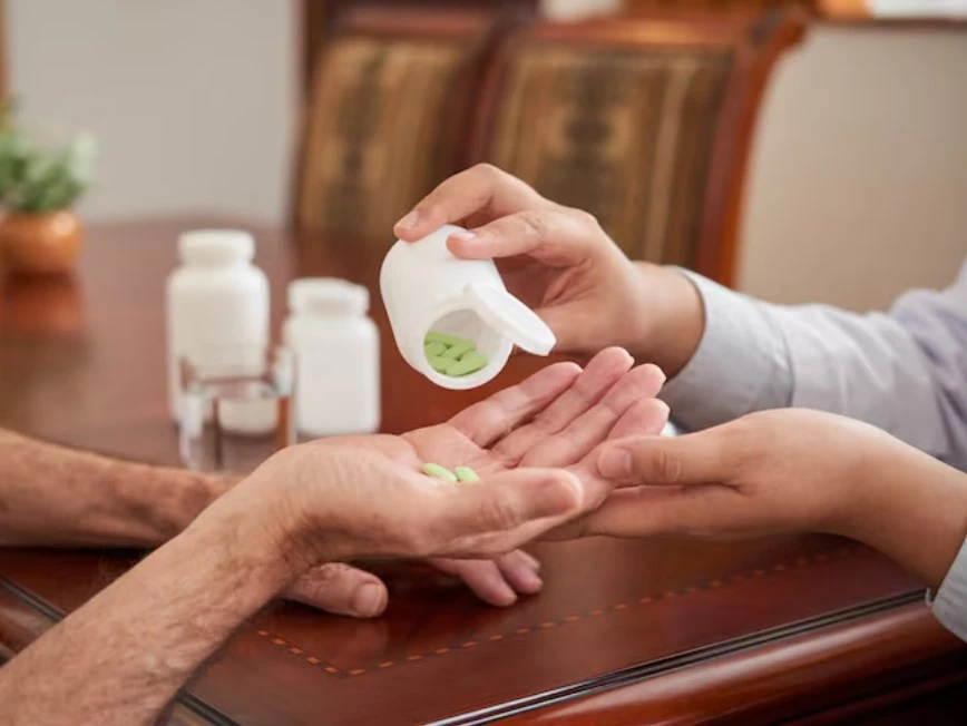 Two people share green pills from a bottle across a table, implying care.