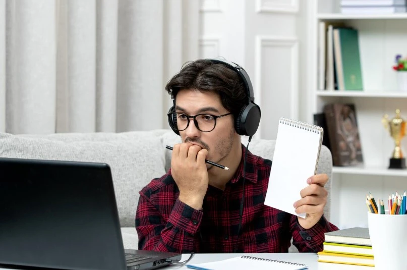 Man with headphones studies laptop, taking notes attentively.
