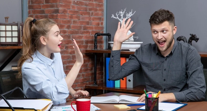 Man and woman high-five at a desk in a lively workspace.