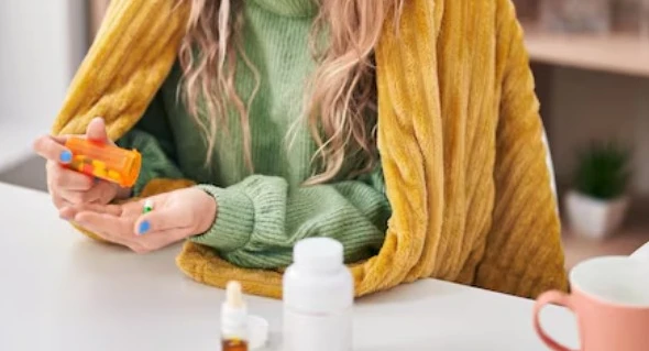 Person in green sweater taking pills at a table with medicines and a mug.