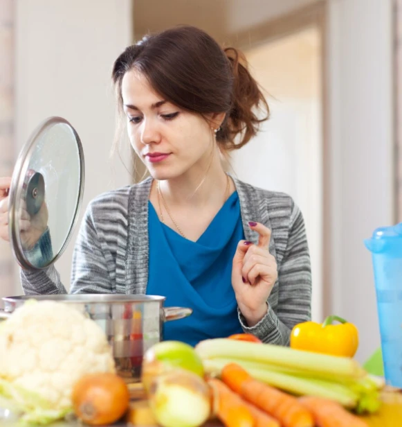 Woman lifting pot lid in kitchen with fresh vegetables on counter