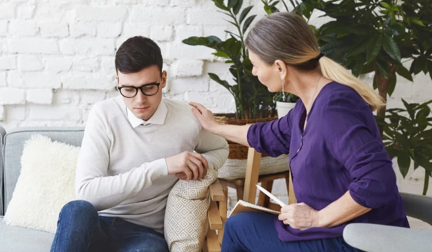 Woman comforts a distressed man on a sofa in a calm room.