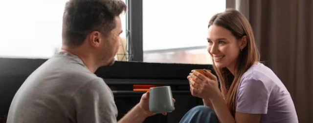 A woman and man smile by a window, holding coffee.